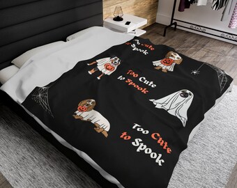 May include: Black and white throw blanket with a spiderweb design and illustrations of dogs dressed as ghosts. The text "Too Cute to Spook" is repeated on the blanket.