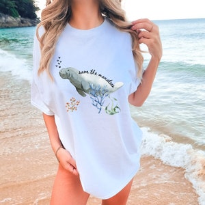 May include: White t-shirt with a watercolor illustration of a manatee swimming in coral. The text "save the manatees" is written in black lettering.