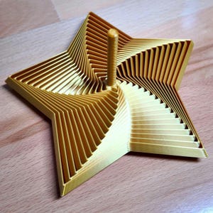 Könnte beinhalten: Ein goldfarbenes, sternförmiges Objekt mit einem zentralen Stift. Das Sterndesign weist geschichtete, abgewinkelte Linien auf, die einen dreidimensionalen Effekt erzeugen. Das Objekt ist wahrscheinlich ein dekoratives Stück oder ein Spielzeug.
