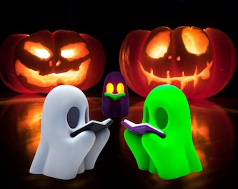 May include: A Halloween-themed image featuring two carved pumpkins with glowing faces and three ghost figurines. One white and one green ghost are reading books, while a purple ghost with yellow eyes is in the background.