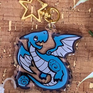 May include: A blue dragon keychain with a gold star-shaped clasp. The dragon has a cartoon design with blue and white details, including wings and a curled tail. The keychain is attached to a gold ring.