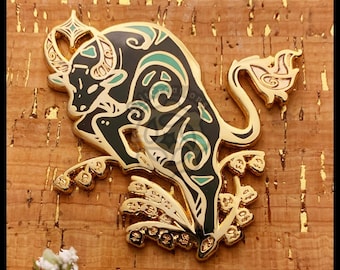 Op de afbeelding: Een goudkleurige metalen pin in de vorm van een stier, met zwarte en teal emaille details. De stier wordt afgebeeld in een dynamische pose, met ingewikkelde patronen. De pin is geplaatst tegen een getextureerde, bruine achtergrond.