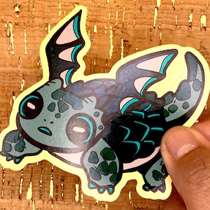 May include: A sticker featuring a cartoon dragon-turtle hybrid with teal, black, and green details. The sticker has a yellow border and is held by a hand. The dragon-turtle has large eyes and wings.
