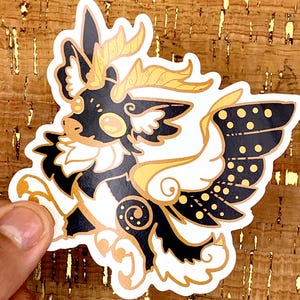 May include: A sticker featuring a stylized creature with black, white, and gold accents. The creature has large wings with black spots, gold horns, and a white chest. The sticker is held by a hand against a textured, brown background.