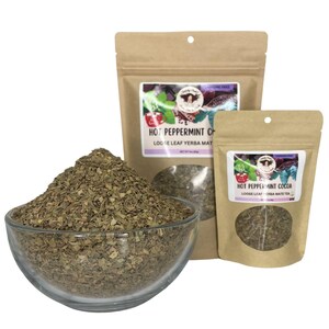 Peut inclure: Un bol en verre transparent rempli de feuilles de th&eacute; en vrac est plac&eacute; devant deux sachets de th&eacute; en papier brun. Les sachets portent les inscriptions "Hot Peppermint Cocoa" et "Hot Peppermint Chai". Les feuilles de th&eacute; sont vertes et brunes.