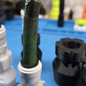K&ouml;nnte beinhalten: Eine Auswahl an 3D-gedruckten Kunststoffteilen in verschiedenen Formen und Farben. Das Bild zeigt wei&szlig;e und schwarze Verbinder, ein gr&uuml;n-schwarzes zylindrisches Bauteil und andere geometrische Formen. Die Teile sind auf einer blauen Oberfl&auml;che angeordnet.