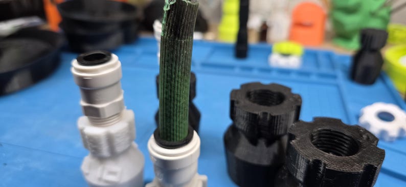 K&ouml;nnte beinhalten: Eine Auswahl an 3D-gedruckten Kunststoffteilen in verschiedenen Formen und Farben. Das Bild zeigt wei&szlig;e und schwarze Verbinder, ein gr&uuml;n-schwarzes zylindrisches Bauteil und andere geometrische Formen. Die Teile sind auf einer blauen Oberfl&auml;che angeordnet.
