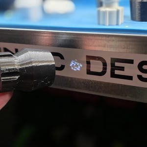 K&ouml;nnte beinhalten: Eine schwarze Taschenlampe mit strukturiertem Griff und einem 3D-gedruckten Aufsatz. Die Taschenlampe wird in der N&auml;he einer Metalloberfl&auml;che mit dem Wort "DESIGN" in schwarzen Buchstaben gehalten. Weitere 3D-gedruckte Objekte sind im Hintergrund zu sehen.