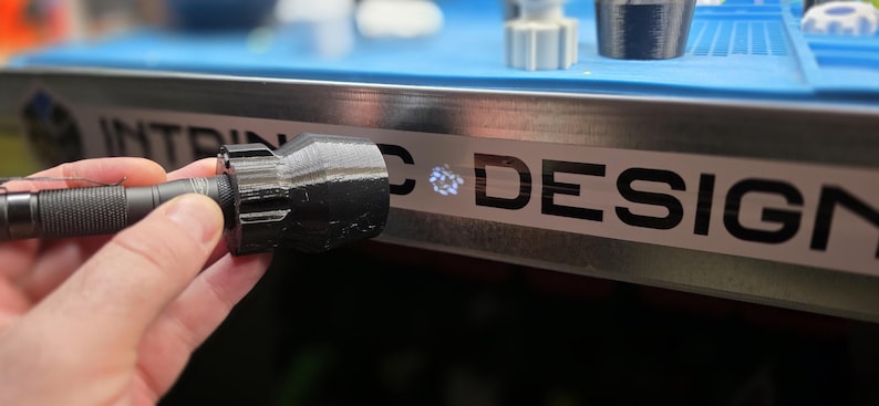 K&ouml;nnte beinhalten: Eine schwarze Taschenlampe mit strukturiertem Griff und einem 3D-gedruckten Aufsatz. Die Taschenlampe wird in der N&auml;he einer Metalloberfl&auml;che mit dem Wort "DESIGN" in schwarzen Buchstaben gehalten. Weitere 3D-gedruckte Objekte sind im Hintergrund zu sehen.