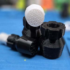 May include: A close-up shot of several black and white 3D-printed components. The components include a cylindrical piece with a textured white top, and several black pieces with various shapes and textures. The items are on a blue surface.
