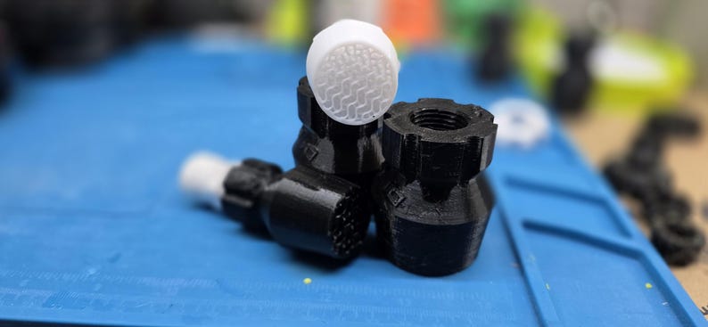 K&ouml;nnte beinhalten: Nahaufnahme mehrerer schwarz-wei&szlig;er 3D-gedruckter Komponenten. Die Komponenten umfassen ein zylindrisches Teil mit einer strukturierten wei&szlig;en Oberseite und mehrere schwarze Teile mit verschiedenen Formen und Texturen. Die Artikel befinden sich auf einer blauen Oberfl&auml;che.