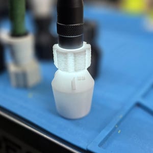 K&ouml;nnte beinhalten: Eine wei&szlig;e Kunststoffkomponente mit einem schwarzen zylindrischen Objekt, das darin eingesetzt ist. Die Komponente hat eine einzigartige Form und befindet sich auf einer blauen Oberfl&auml;che. Weitere &auml;hnliche Komponenten sind im Hintergrund sichtbar.