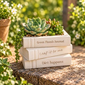 May include: A stack of three off-white rectangular planters shaped like books, with the text Green Thumb Journal, Leaf it to me, and Dirt happens engraved on the spines. The top planter holds a variety of green succulents and trailing beads.