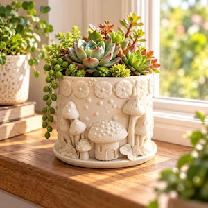 May include: A cylindrical cream-colored ceramic planter featuring embossed mushroom and floral motifs, filled with a variety of green and reddish succulents, including a trailing string of pearls plant, resting on a matching circular saucer.