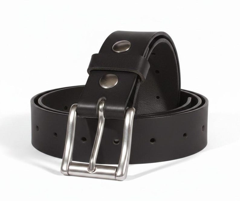 May include: A black leather belt with a silver-colored buckle. The belt is rolled up, showing the buckle and the belt's holes. The belt has two silver rivets near the buckle. The background is white.