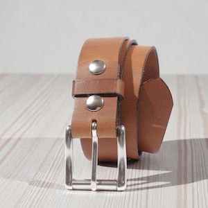 May include: A rolled-up, light brown leather belt with a silver-colored buckle and two silver rivets. The belt is on a light-colored wooden surface.