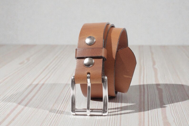 May include: A rolled-up, light brown leather belt with a silver-colored buckle and two silver rivets. The belt is on a light-colored wooden surface.