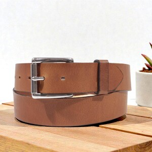 May include: A brown leather belt with a silver buckle is displayed on a wooden surface. The belt is rolled up, showcasing its texture and design. A small succulent plant in a white pot is in the background.
