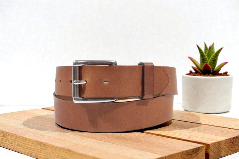 May include: A brown leather belt with a silver buckle is displayed on a wooden surface. The belt is rolled up, showcasing its texture and design. A small succulent plant in a white pot is in the background.