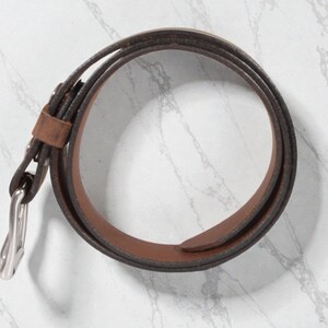 May include: A rolled brown leather belt with a silver-colored buckle. The belt is a medium brown color with a darker brown edge. The buckle is a simple, classic design.
