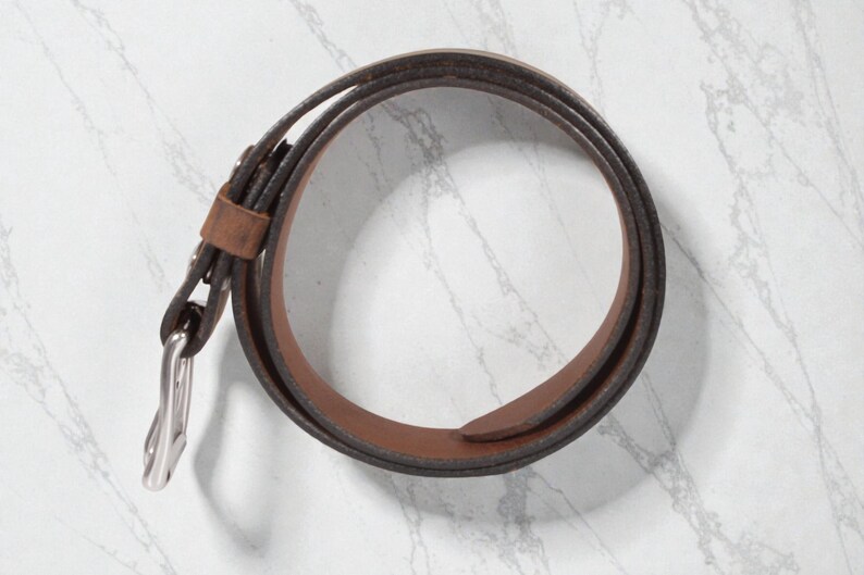 May include: A rolled brown leather belt with a silver-colored buckle. The belt is a medium brown color with a darker brown edge. The buckle is a simple, classic design.