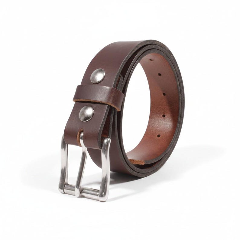 May include: A brown leather belt with a silver-colored buckle and two silver rivets. The belt is rolled up, showcasing its rich color and texture. A classic accessory.