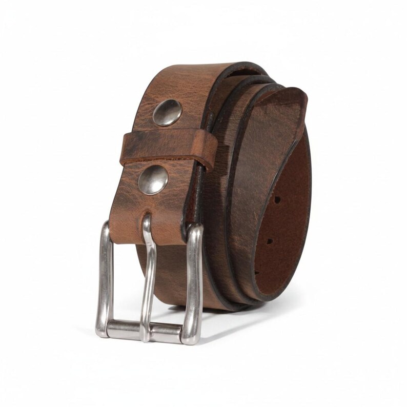May include: A rolled-up brown leather belt with a silver-colored buckle and two silver rivets. The belt has a distressed appearance, suggesting a vintage or worn look. The background is plain white.