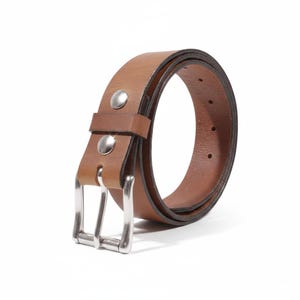 May include: A brown leather belt with a silver-colored buckle and two silver rivets. The belt is curved, showing the inside and outside of the leather. The belt is likely 1.25 inches wide.