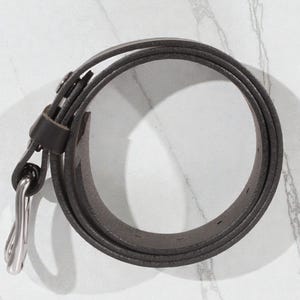 May include: A rolled-up, dark gray leather belt with a silver-colored buckle. The belt is thin and appears to be made of high-quality leather. The buckle is a simple, classic design. The belt is on a white marble surface.