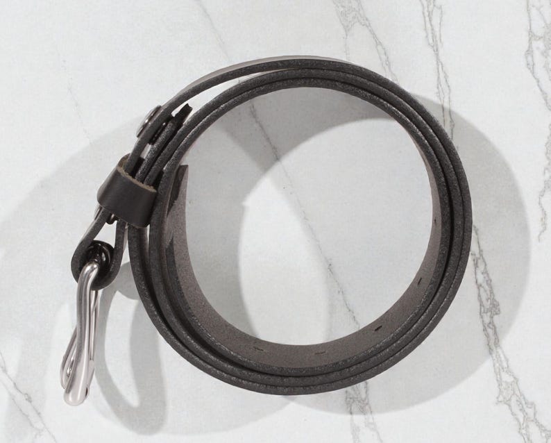 May include: A rolled-up, dark gray leather belt with a silver-colored buckle. The belt is thin and appears to be made of high-quality leather. The buckle is a simple, classic design. The belt is on a white marble surface.