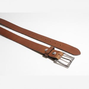 May include: A brown leather belt with a silver-colored buckle. The belt has several holes for adjustment and is made of a smooth, solid material. The buckle is rectangular and has a classic design.