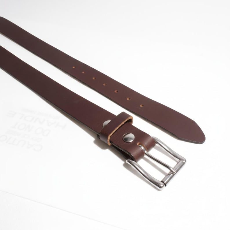 May include: A brown leather belt with a silver-colored buckle. The belt has a rectangular buckle and several holes for adjustment. The leather is a rich, dark brown color.