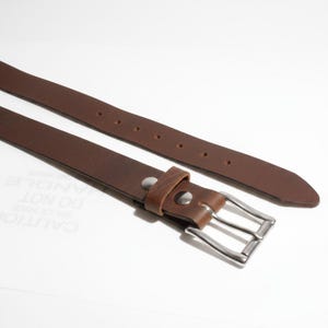 May include: A brown leather belt with a silver-colored buckle. The belt has several holes for adjustment and is displayed against a white background. The leather appears to be textured.