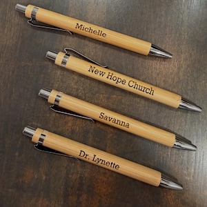 May include: Four bamboo pens with silver accents and black ink. Each pen is engraved with a name or phrase: "Michelle", "New Hope Church", "Savanna", and "Dr. Lynette". The pens are arranged on a dark wooden surface.