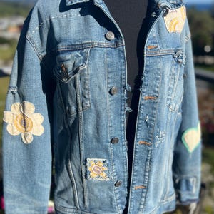 May include: Light blue denim jacket with decorative patches. Patches include a flower, a rainbow, and a heart. The jacket has button closures, pockets, and distressed details. The jacket is displayed on a black mannequin.