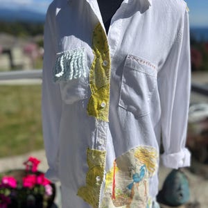 May include: White button-down shirt with long sleeves, featuring patchwork details. The shirt has yellow and light blue accents, with a whimsical illustration on the front. The shirt has a pocket on the chest and is displayed on a mannequin.