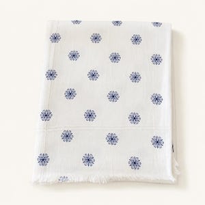 May include: A white rectangular textile featuring a repeating pattern of small, dark blue, multi-petaled flower motifs arranged in a grid. The fabric has a visible horizontal seam across the upper section and a frayed, fringed edge at the bottom.
