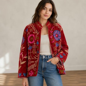 May include: A deep red velvet jacket featuring vibrant multi-colored floral and heart-shaped embroidery in shades of pink, blue, and gold. The jacket has a high collar, long sleeves, and a straight hemline, worn over a white tank top and blue jeans.
