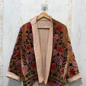 May include: A brown velvet jacket featuring colorful floral embroidery in red, pink, and purple with black vine patterns. The jacket has an open front, long sleeves with folded cuffs, and a light-colored interior lining with thin vertical stripes.
