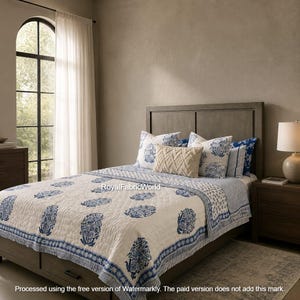 May include: A cream-colored quilted bedspread featuring a repeating blue floral block print pattern. The bedding set includes several decorative pillows in matching blue and white floral designs, along with a solid textured cream-colored pillow.