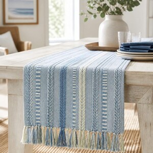 May include: A rectangular table runner featuring a woven pattern of vertical stripes in shades of light blue, medium blue, and cream. The runner is finished with long, knotted fringe tassels in cream and light blue along the bottom edge.