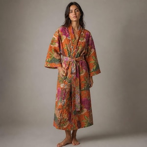 May include: A long-sleeved, calf-length robe featuring a vibrant orange base with a multi-colored floral and botanical print. The garment includes a matching fabric tie belt at the waist, wide sleeves, and visible quilted stitching throughout the fabric.