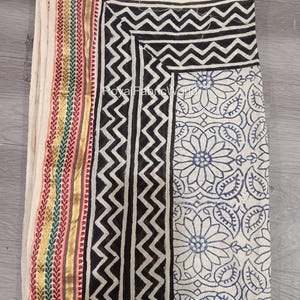May include: A folded textile featuring a cream-colored base with various patterns. The design includes black zigzag and floral prints, along with a striped border in gold, red, green, and yellow. The text "Royal Fabric World" is visible.