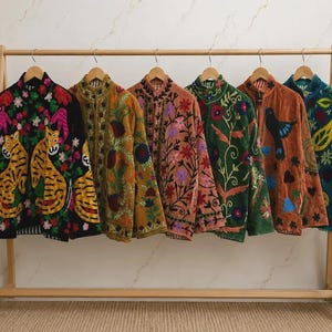 May include: Six long-sleeved jackets hanging on a wooden rack, each featuring distinct colorful embroidery. The designs include tigers, floral patterns, birds, and paisley motifs in various colors like black, mustard, pink, green, and blue.