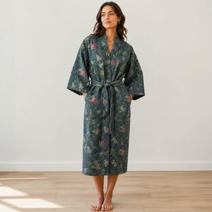 May include: A long-sleeved, calf-length robe in a dark teal-grey fabric featuring a multi-colored floral pattern. The garment includes a matching fabric tie belt at the waist, a V-neckline, and wide sleeves, showcasing visible quilted stitching throughout.
