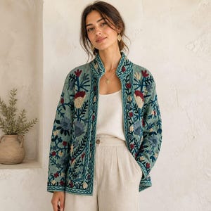 May include: A light blue long-sleeved jacket featuring intricate floral embroidery in shades of red, cream, and dark blue. The jacket has a stand-up collar, an open front design, and is worn over a plain white tank top and light beige trousers.