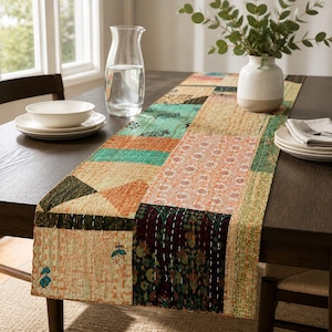 May include: A rectangular table runner featuring a patchwork design of various fabric squares in shades of orange, teal, green, and cream. The runner is decorated with intricate white kantha stitching and floral patterns, laid flat across a dark wooden table.