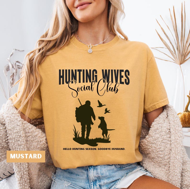 K&ouml;nnte beinhalten: Ein senfgelbes T-Shirt mit dem Text "Hunting Wives Social Club" in Schwarz. Das Shirt zeigt eine Silhouette eines J&auml;gers mit einem Hund und V&ouml;geln, die &uuml;ber ihm fliegen. Der Text "Hello Hunting Season. Goodbye Husband." ist unter dem Bild gedruckt.
