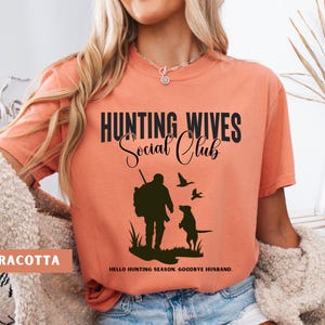 K&ouml;nnte beinhalten: Ein terracottafarbenes T-Shirt mit dem Text "Hunting Wives Social Club" in Schwarz. Das Shirt zeigt eine Silhouette eines J&auml;gers mit einem Hund und V&ouml;geln. Der Text "Hello hunting season. Goodbye husband." ist unter der Grafik gedruckt.