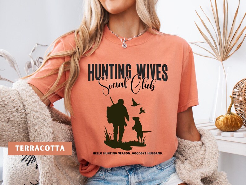 K&ouml;nnte beinhalten: Ein terracottafarbenes T-Shirt mit dem Text "Hunting Wives Social Club" in Schwarz. Das Shirt zeigt eine Silhouette eines J&auml;gers mit einem Hund und V&ouml;geln. Der Text "Hello hunting season. Goodbye husband." ist unter der Grafik gedruckt.